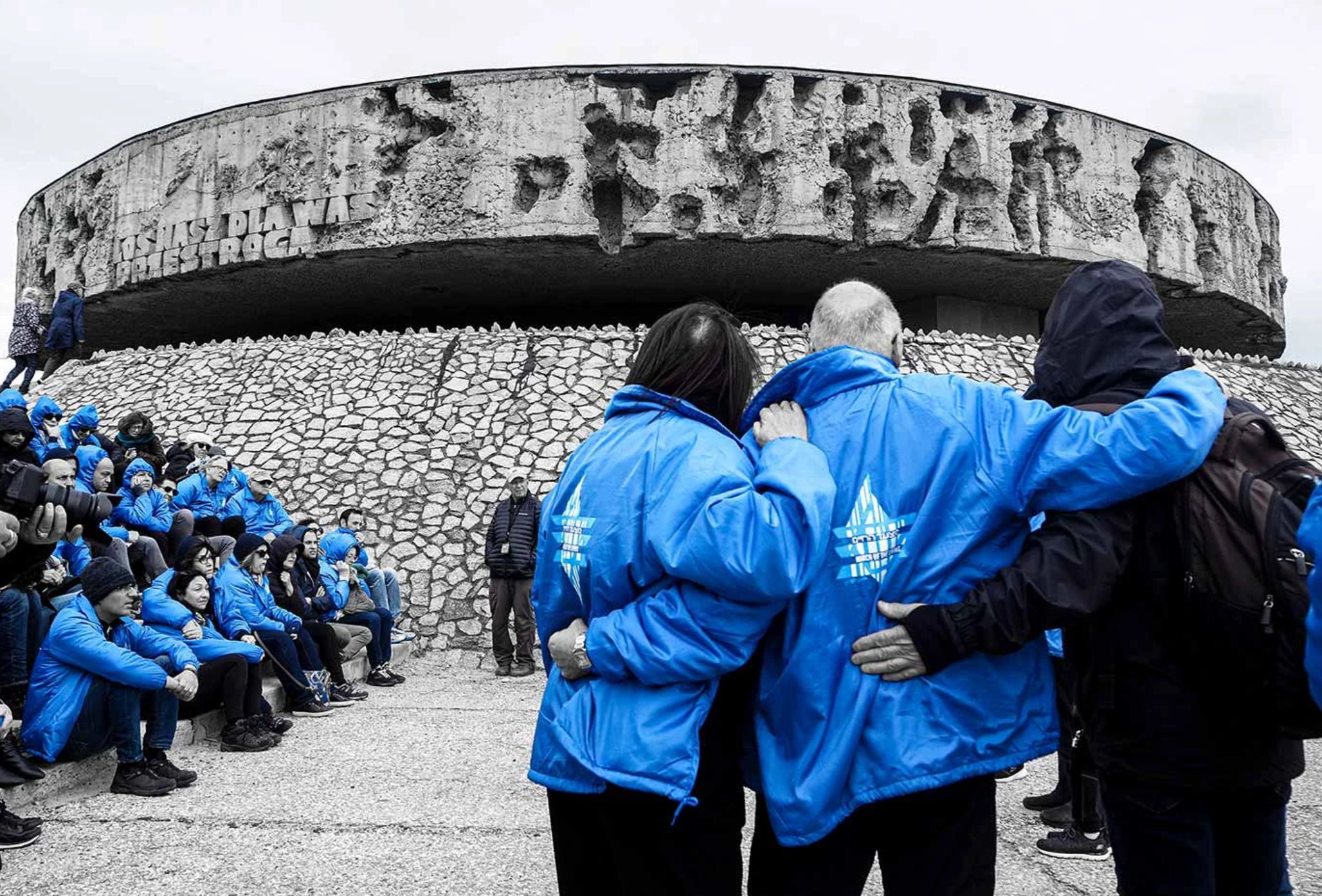 People in blue jackets in an otherwise black and white photograph. They are standing with arms around each other in front of a large memorial with a Polish motto.