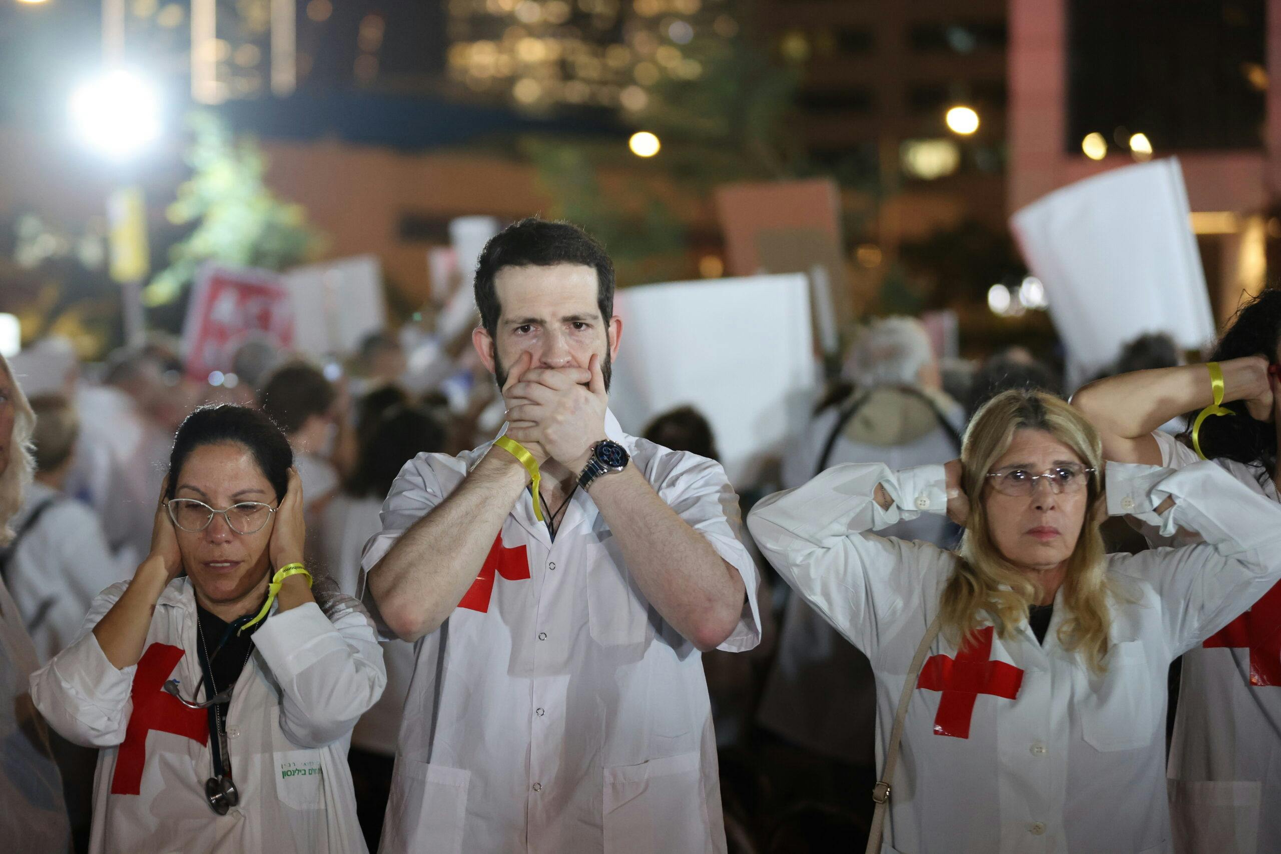 3 people in white coats with red crosses on them, one with hands over mouth, one with hands over ears, one with hands over eyes.
