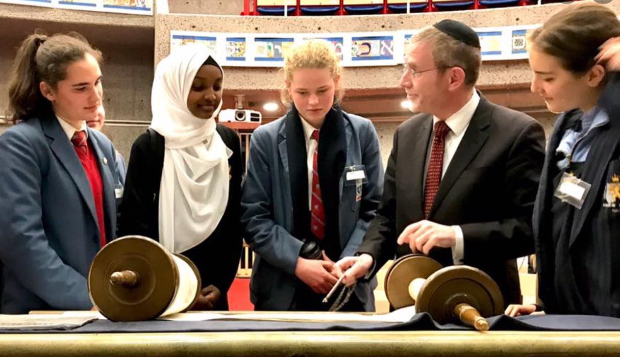 Teacher wearing a Jewish skullcap with students, including one in a hijab, looking at a Torah scroll