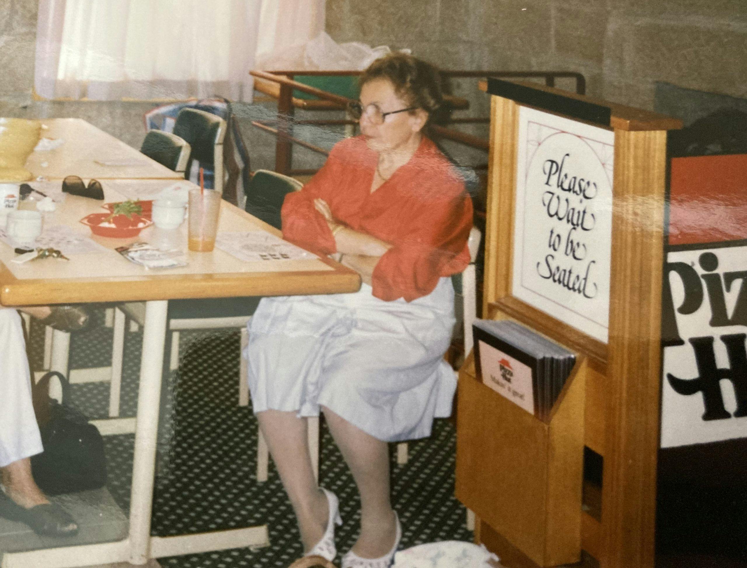 Older woman sitting at Pizza Hut with a red top and white skirt, arms folded